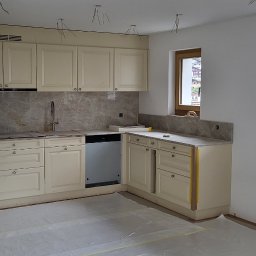 Remonty - Jasna, klasyczna kuchnia w trakcie montażu, z widocznymi kablami zwisającymi z sufitu i zabezpieczoną folią podłogą, okno z widokiem na zabudowę górską.