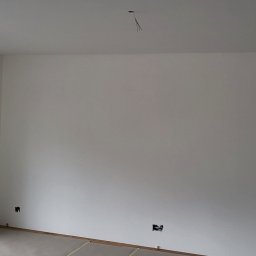 Remonty - Wnętrze pokoju po świeżym malowaniu ścian na biało, widoczne gniazdka elektryczne bez osprzętu oraz wystające przewody z sufitu, z drewnianą ramą okienną.