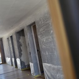 Remonty - Wnętrze w trakcie prac wykończeniowych, ściany i sufit po nałożeniu gładzi, zabezpieczona podłoga, widoczne ościeżnice drzwiowe.