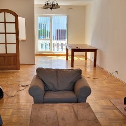 Remonty - Jasne wnętrze salonu z parkietem w jodełkę, widoczne meble wypoczynkowe, drewniany stół, kominek z cegły oraz drzwi balkonowe z kratą zabezpieczającą.