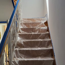 Remonty - Schody drewniane zabezpieczone folią malarską przed malowaniem ścian, widoczne taśmy malarskie na krawędziach, światło dzienne wpadające przez okno dachowe.