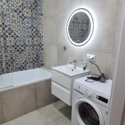 Remonty - Wykończona łazienka z mozaiką nad wanną, okrągłym lustrem z podświetleniem LED, białą umywalką z szafką i pralką Electrolux z akcesoriami na górze.