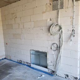 Remonty - Surowy stan wnętrza z białymi bloczkami, widoczne kable elektryczne zwisające ze ściany, niebieska rura instalacyjna przy podłodze i otwór na rozdzielnicę elektryczną.