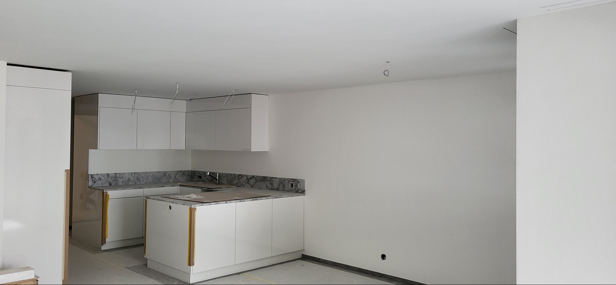 Wnętrze w trakcie wykańczania: biała zabudowa kuchenna z szarym blatem z efektem marmuru, widoczne przewody elektryczne zwisające z sufitu, surowe ściany pomalowane na biało.