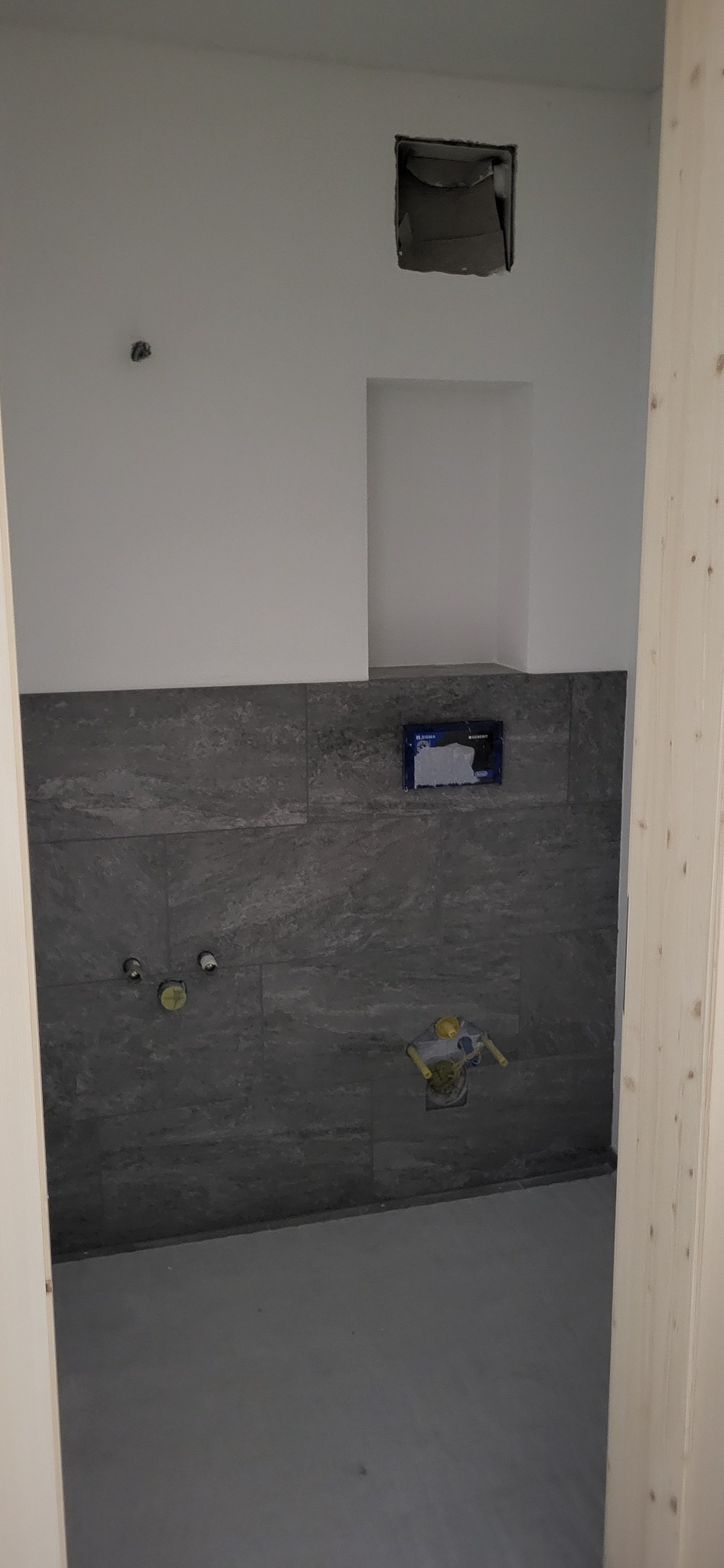 Wnętrze łazienki w trakcie wykańczania w Nowym Dworze Mazowieckim, widoczne szare płytki na ścianie, przygotowane instalacje wodne i otwór w ścianie nad stelażem WC.