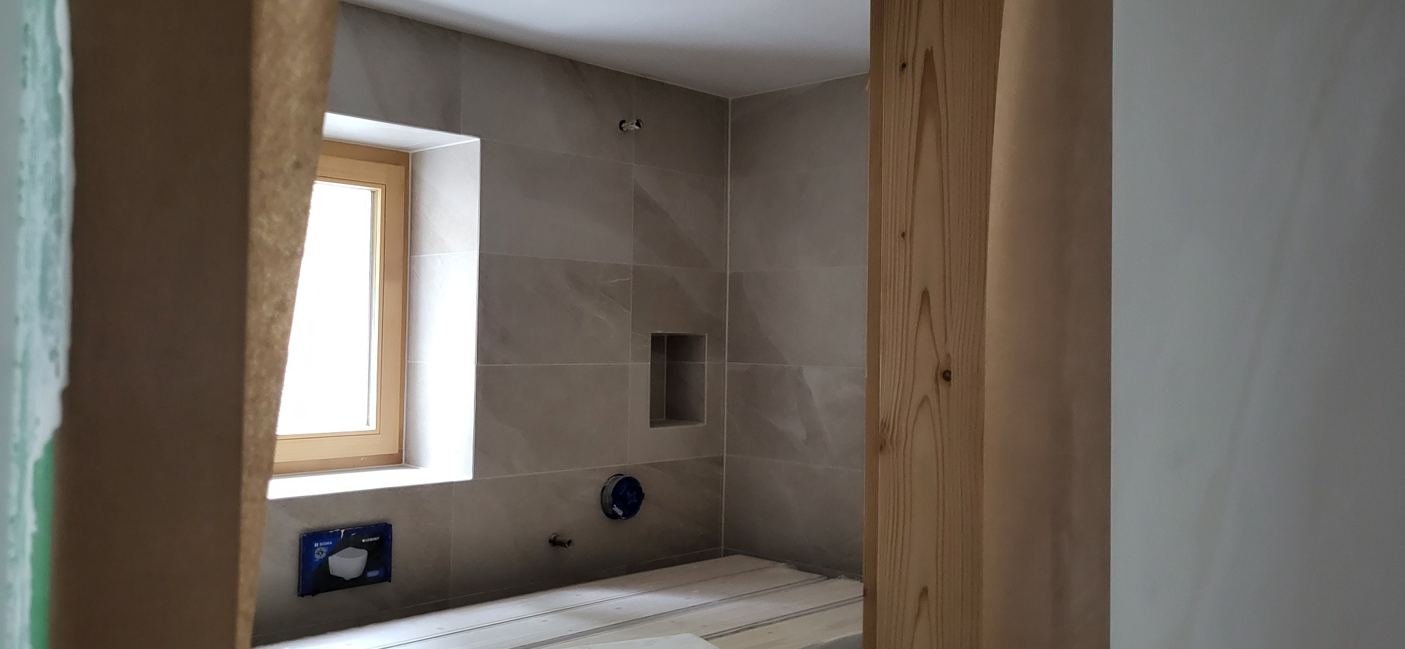 Łazienka w trakcie wykończenia: szare płytki ceramiczne na ścianach, drewniana rama okienna z widokiem na zewnątrz, podłoga przygotowana pod ułożenie płytek, widoczne elementy instalacji podtynkowych.