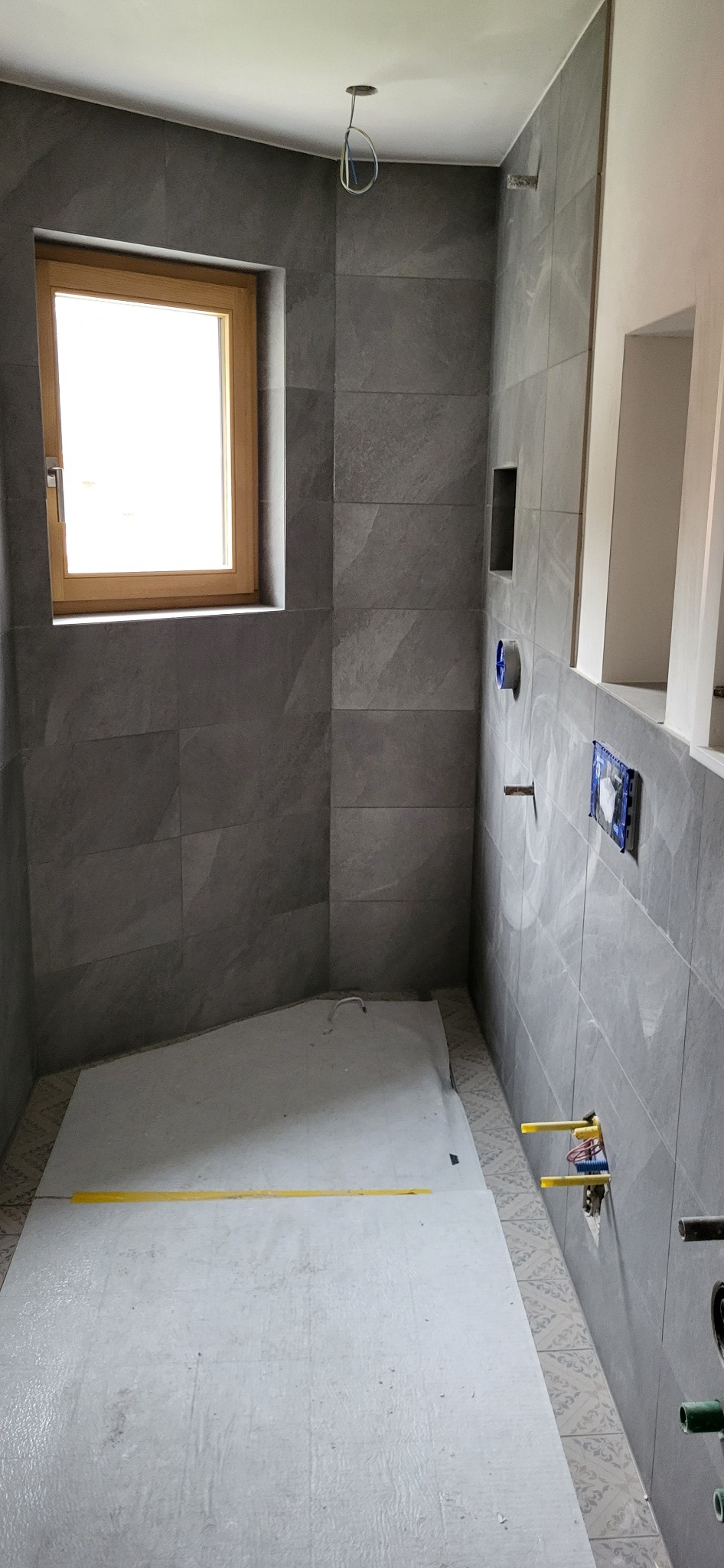 Szara łazienka w trakcie prac wykończeniowych: ułożone płytki imitujące kamień, zabezpieczona podłoga, widoczne instalacje wodno-kanalizacyjne i otwór pod oświetlenie.