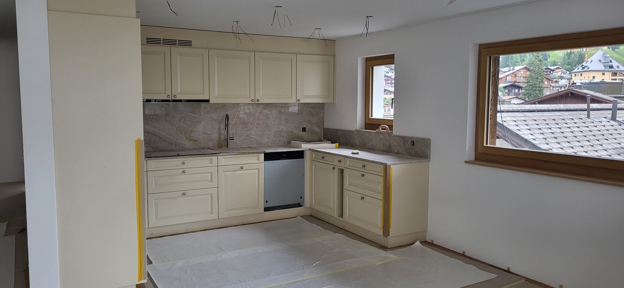 Jasna, klasyczna kuchnia w trakcie montażu, z widocznymi kablami zwisającymi z sufitu i zabezpieczoną folią podłogą, okno z widokiem na zabudowę górską.