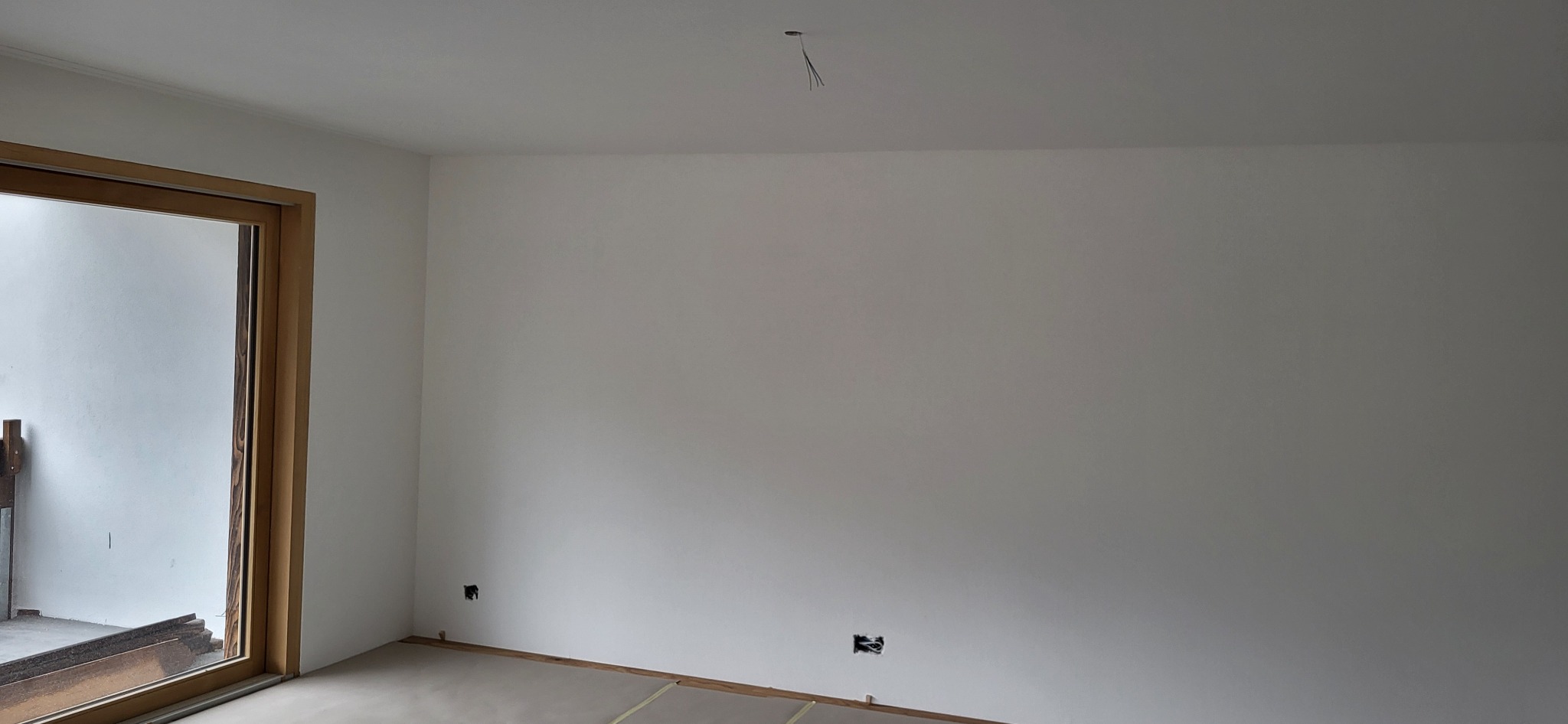 Wnętrze pokoju po świeżym malowaniu ścian na biało, widoczne gniazdka elektryczne bez osprzętu oraz wystające przewody z sufitu, z drewnianą ramą okienną.