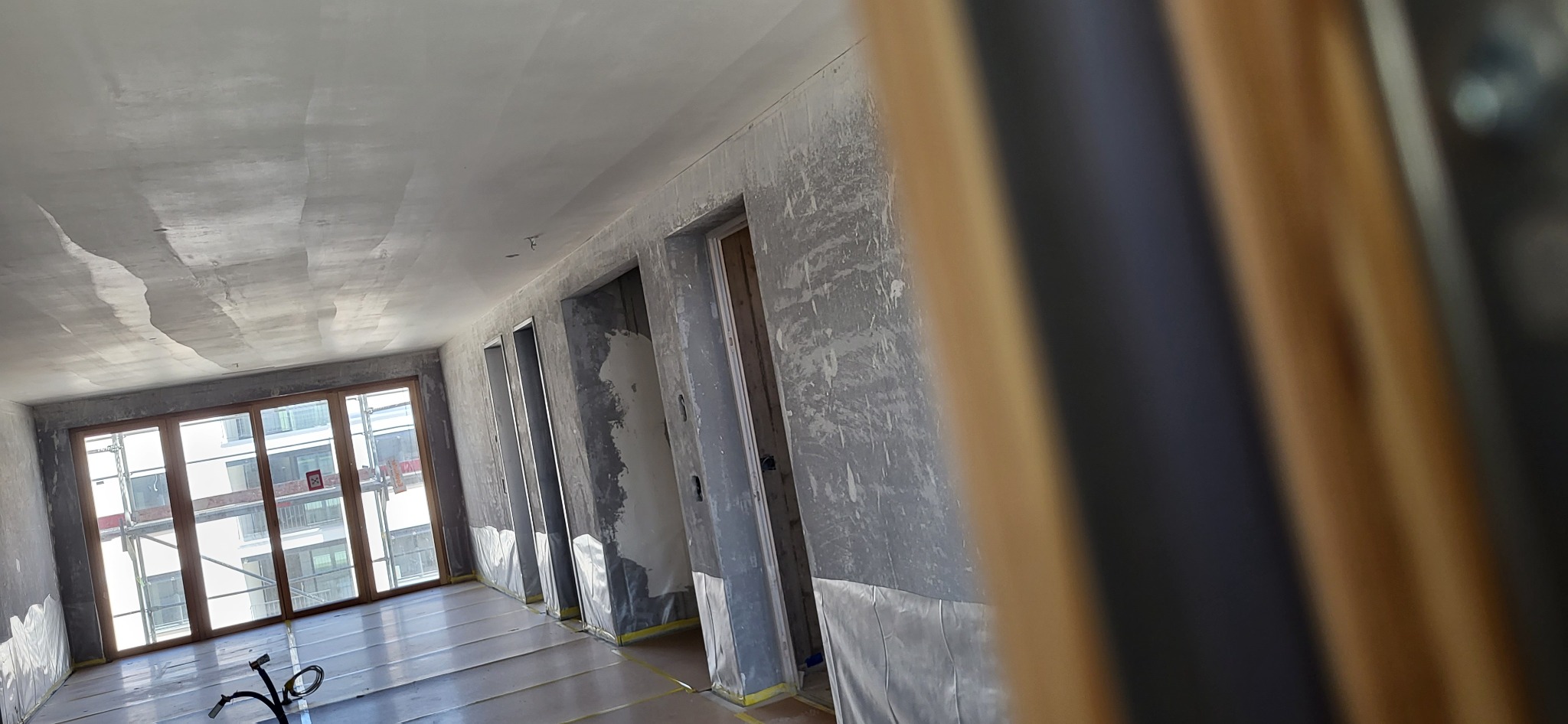 Wnętrze w trakcie prac wykończeniowych, ściany i sufit po nałożeniu gładzi, zabezpieczona podłoga, widoczne ościeżnice drzwiowe.