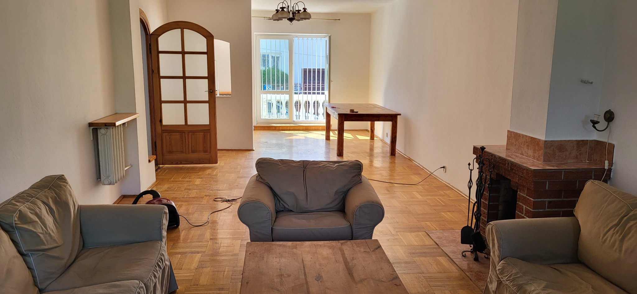 Jasne wnętrze salonu z parkietem w jodełkę, widoczne meble wypoczynkowe, drewniany stół, kominek z cegły oraz drzwi balkonowe z kratą zabezpieczającą.