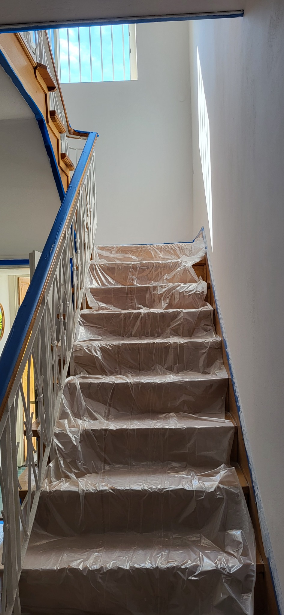 Schody drewniane zabezpieczone folią malarską przed malowaniem ścian, widoczne taśmy malarskie na krawędziach, światło dzienne wpadające przez okno dachowe.