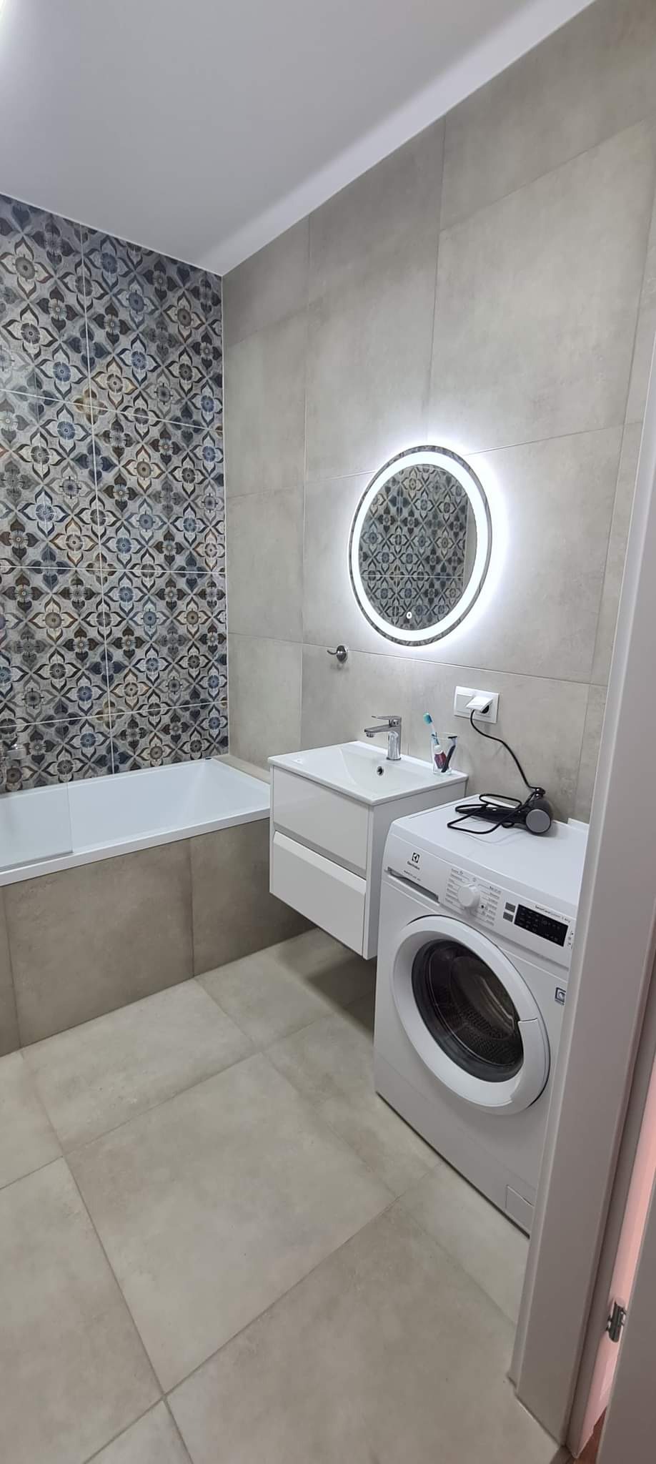 Wykończona łazienka z mozaiką nad wanną, okrągłym lustrem z podświetleniem LED, białą umywalką z szafką i pralką Electrolux z akcesoriami na górze.