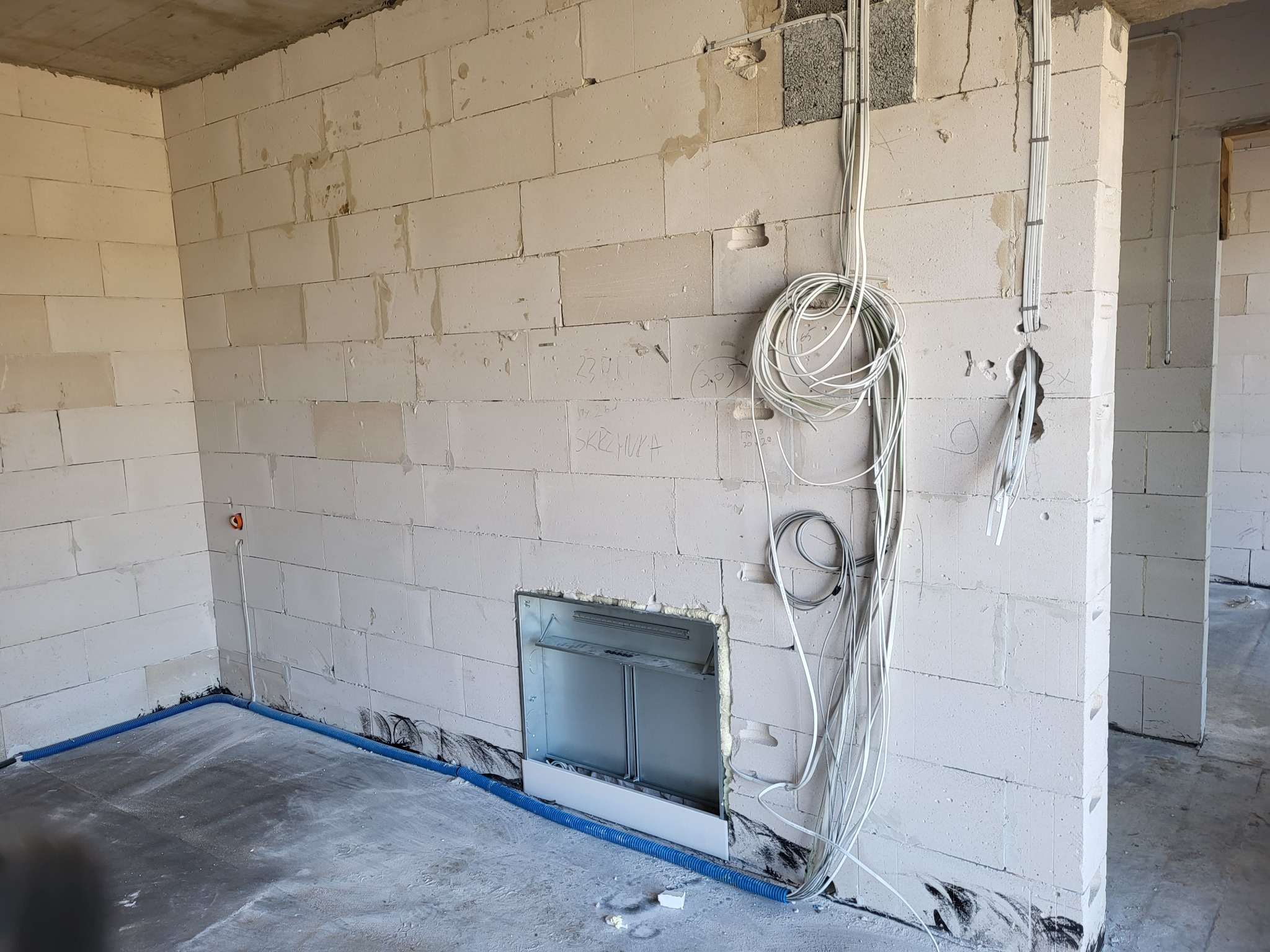 Surowy stan wnętrza z białymi bloczkami, widoczne kable elektryczne zwisające ze ściany, niebieska rura instalacyjna przy podłodze i otwór na rozdzielnicę elektryczną.
