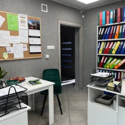 R.G Partners Sp. z o.o - Wnętrze biura z regałem pełnym kolorowych segregatorów, biurkiem z jabłkami i zielonym krzesłem welurowym, na ścianie tablica korkowa z dokumentami i kalendarzem.