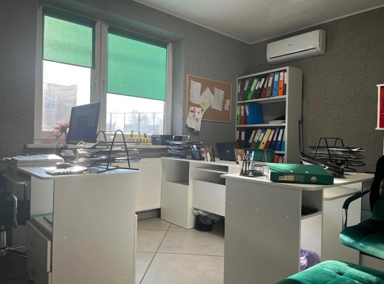 Biuro z dwoma białymi biurkami, komputerem, segregatorami, tablicą korkową z notatkami, zielonymi roletami i klimatyzacją na szarej ścianie, widok z perspektywy krzesła.