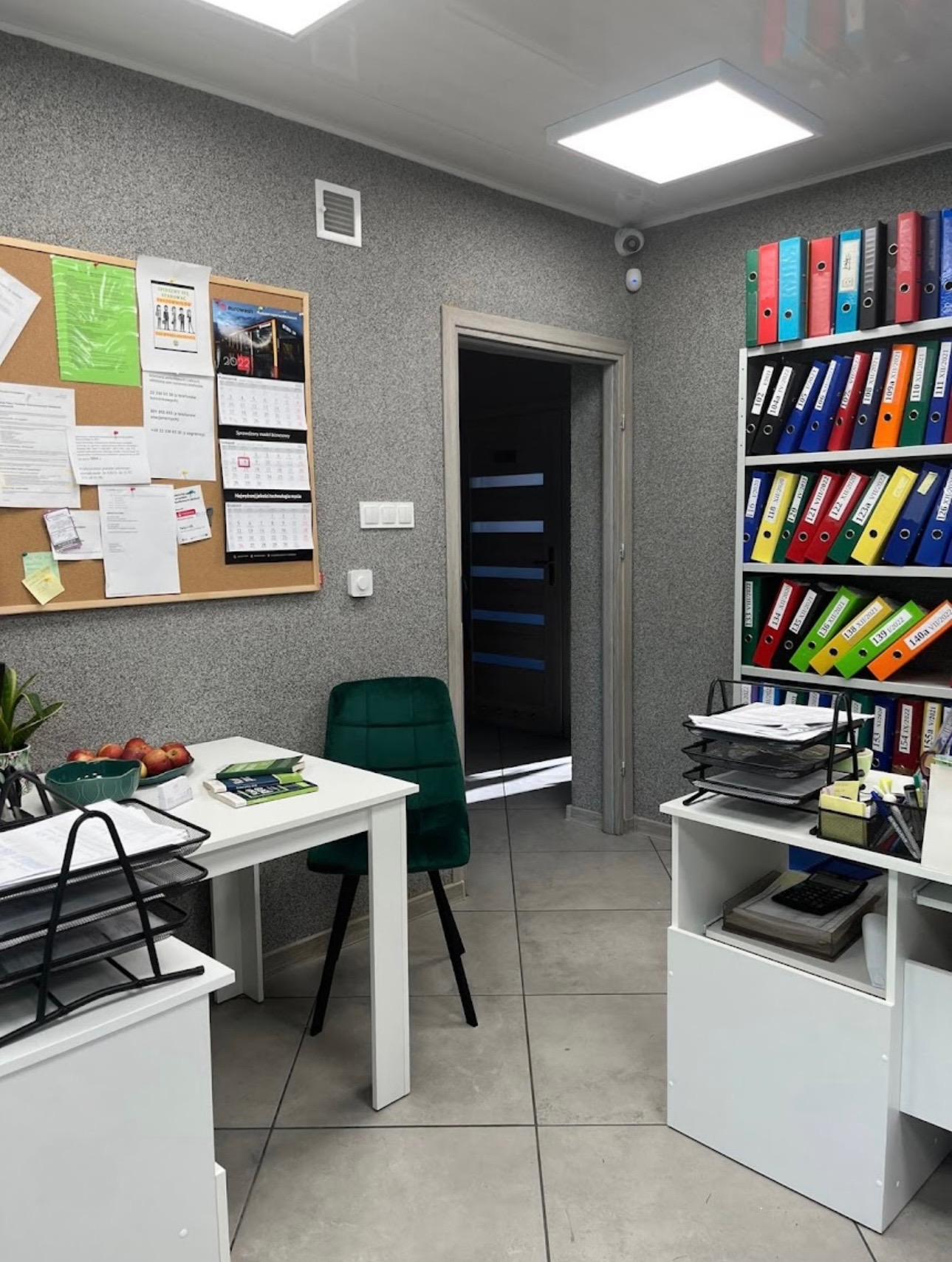 Wnętrze biura z regałem pełnym kolorowych segregatorów, biurkiem z jabłkami i zielonym krzesłem welurowym, na ścianie tablica korkowa z dokumentami i kalendarzem.