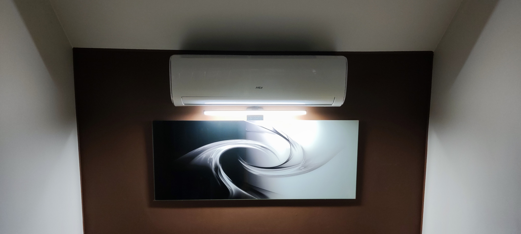 Biały klimatyzator ścienny marki MDJ zamontowany nad podświetlonym obrazem abstrakcyjnym na brązowej ścianie w pokoju.
