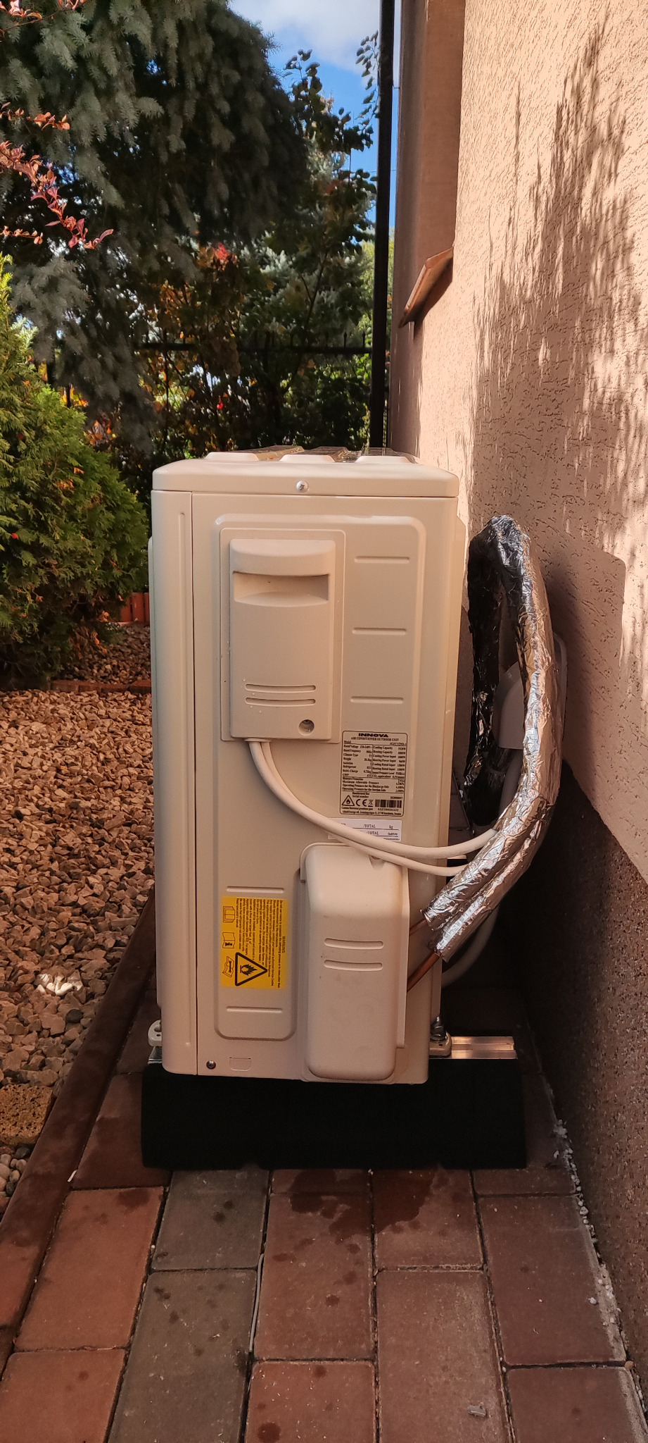 Zewnętrzna jednostka klimatyzacji zamontowana przy ścianie budynku, widoczne rury w izolacji termicznej, etykieta z ostrzeżeniem i specyfikacją techniczną.