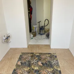 Fragment łazienki w trakcie remontu z ułożoną częścią podłogi z płytek imitujących marmur oraz płytek z motywem roślinnym; w tle widoczny odkurzacz, worek z zaprawą i otwarte drzwi.