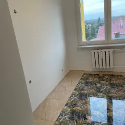 Pokój po remoncie z częściowo ułożonymi płytkami podłogowymi w dwa różne wzory: beżowe imitujące kamień i wzorzyste w motywy roślinne, widoczne otwory w ścianie pod gniazdka elektryczne, biały...