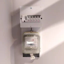 Zamontowany licznik elektryczny typu A52 wraz z rozdzielnicą z bezpiecznikami topikowymi Etimats B16, na białej ścianie z widocznym cieniem.
