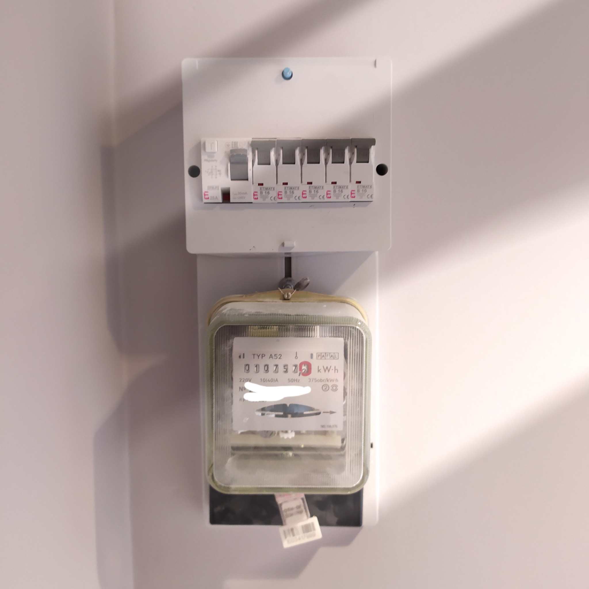 Zamontowany licznik elektryczny typu A52 wraz z rozdzielnicą z bezpiecznikami topikowymi Etimats B16, na białej ścianie z widocznym cieniem.