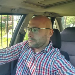 Mężczyzna w okularach i koszuli w kratę siedzi w samochodzie, widok z wnętrza pojazdu na zieleń za oknem.