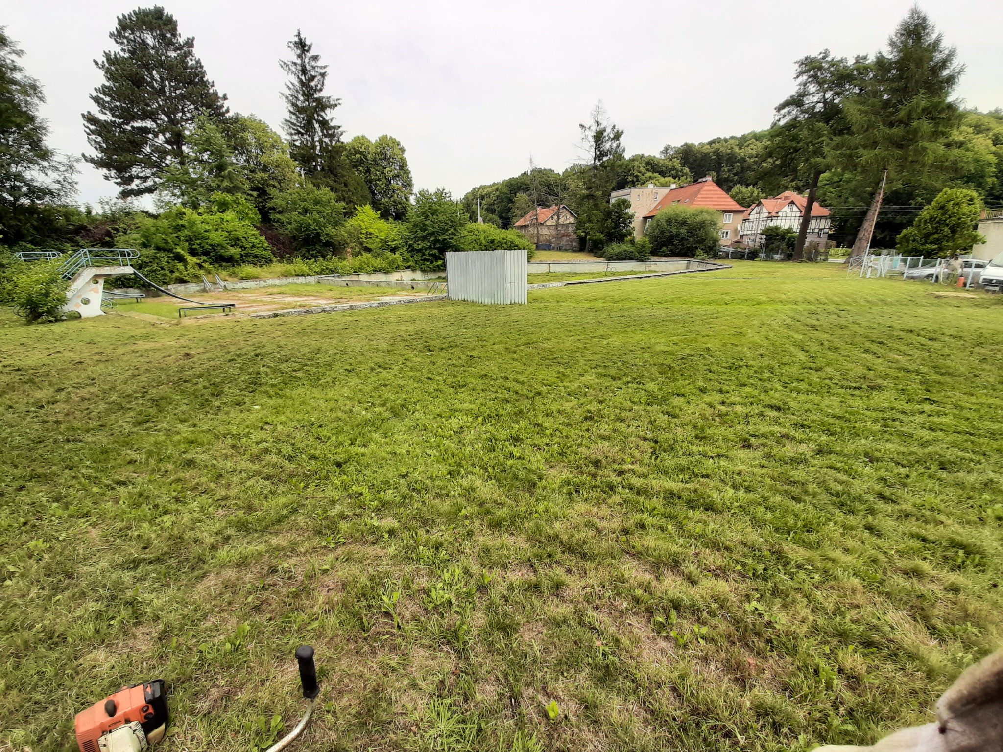 Szeroki kadr na częściowo skoszoną trawę na terenie dawnego kąpieliska, w tle budynki mieszkalne i drzewa, w dolnym rogu widoczna kosa spalinowa.
