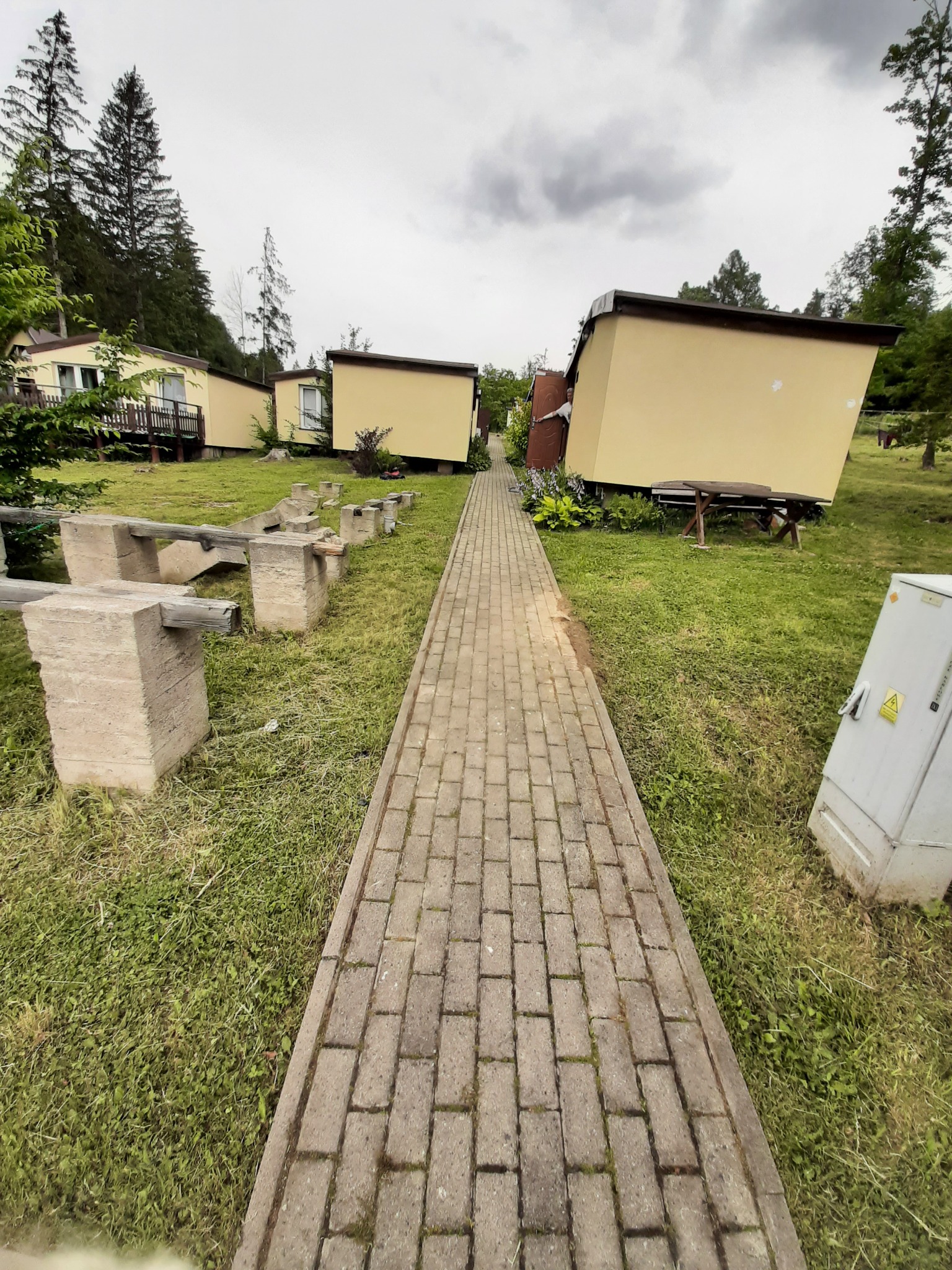 Długa, brukowana ścieżka prowadzi przez trawnik między żółtymi domkami letniskowymi z drewnianymi elementami, widoczne betonowe bloki i drewniane belki na trawie.