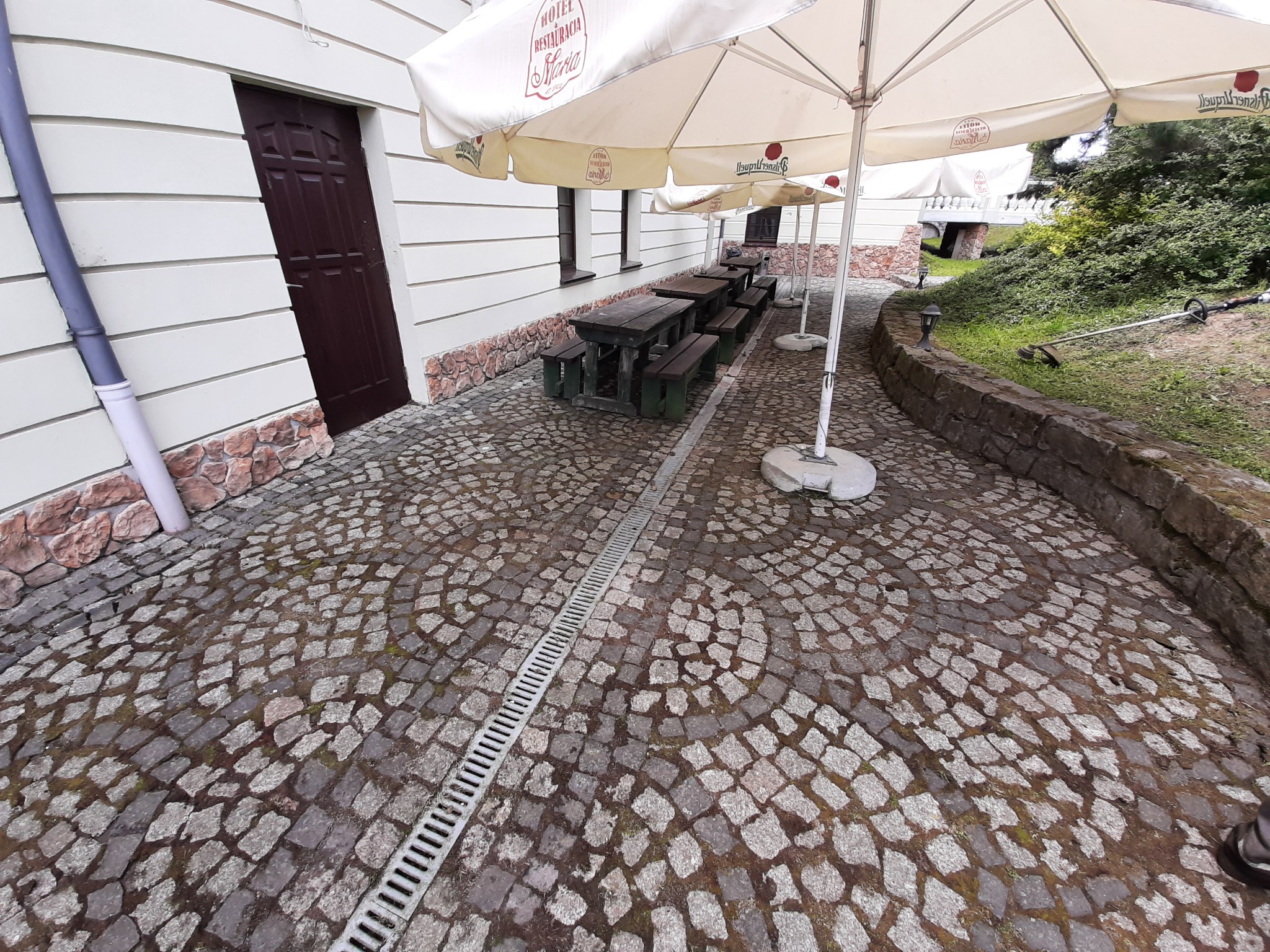 Ułożona z kostki brukowej ścieżka przed hotelem z parasolami, stolikami i ławkami, w tle widoczny fragment trawnika z podkaszarką.