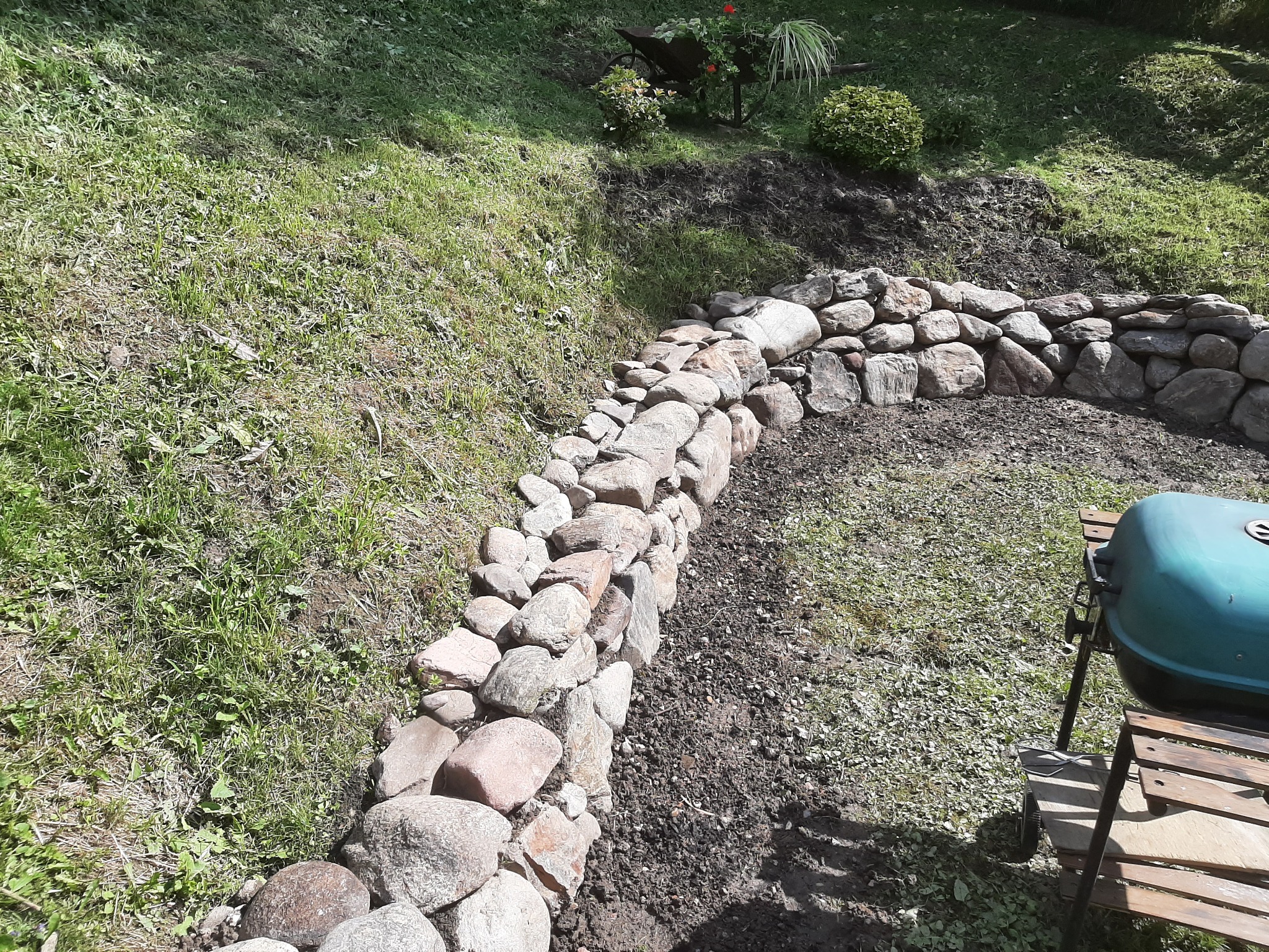 Kamienny murek oporowy oddzielający trawnik od rabaty, z widoczną taczką z kwiatami w tle, na nierównym terenie.