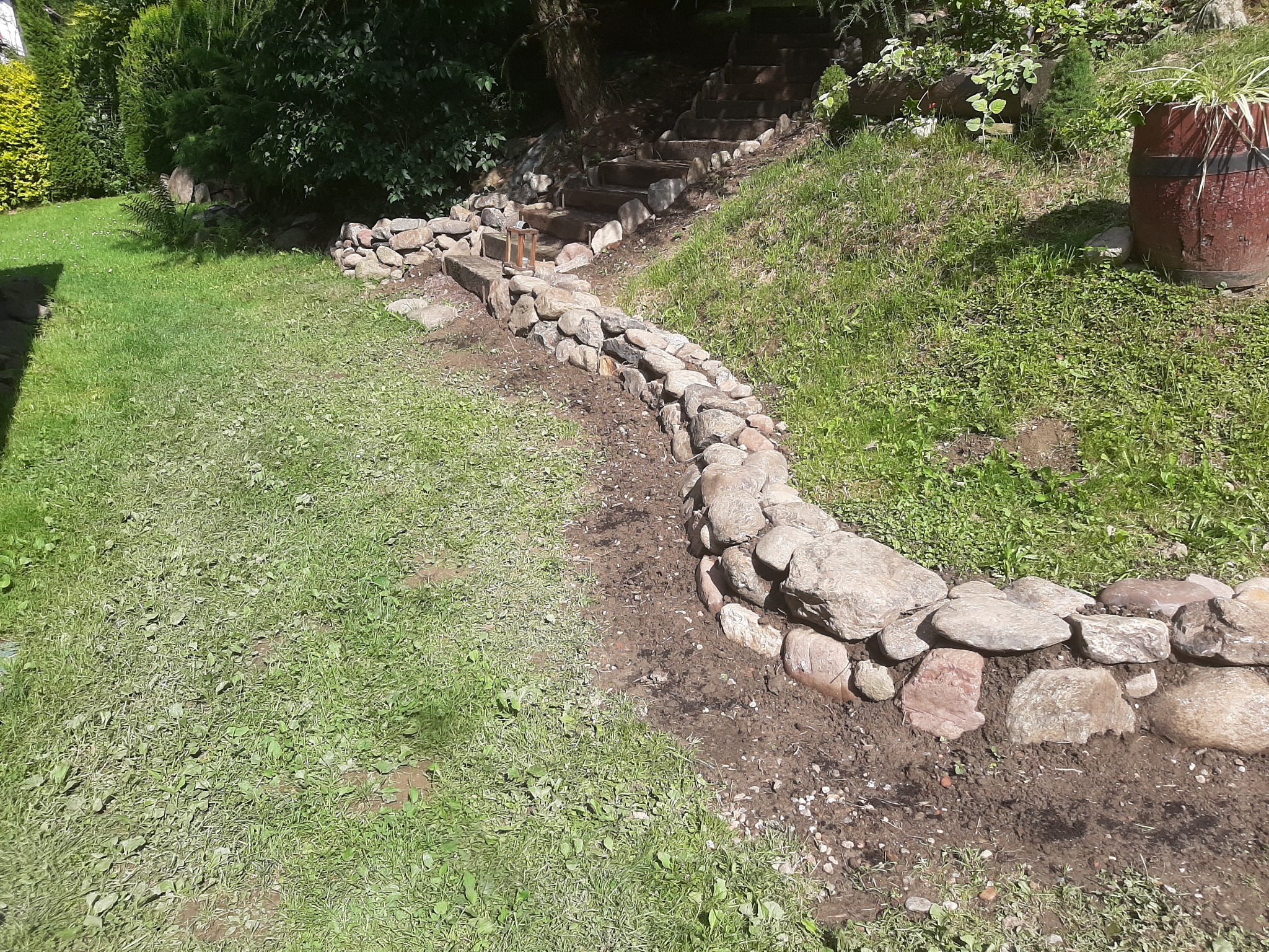Ułożony z kamieni brzeg rabaty w ogrodzie, prowadzący w górę zbocza obok drewnianych schodów, z widoczną ziemią i trawą.