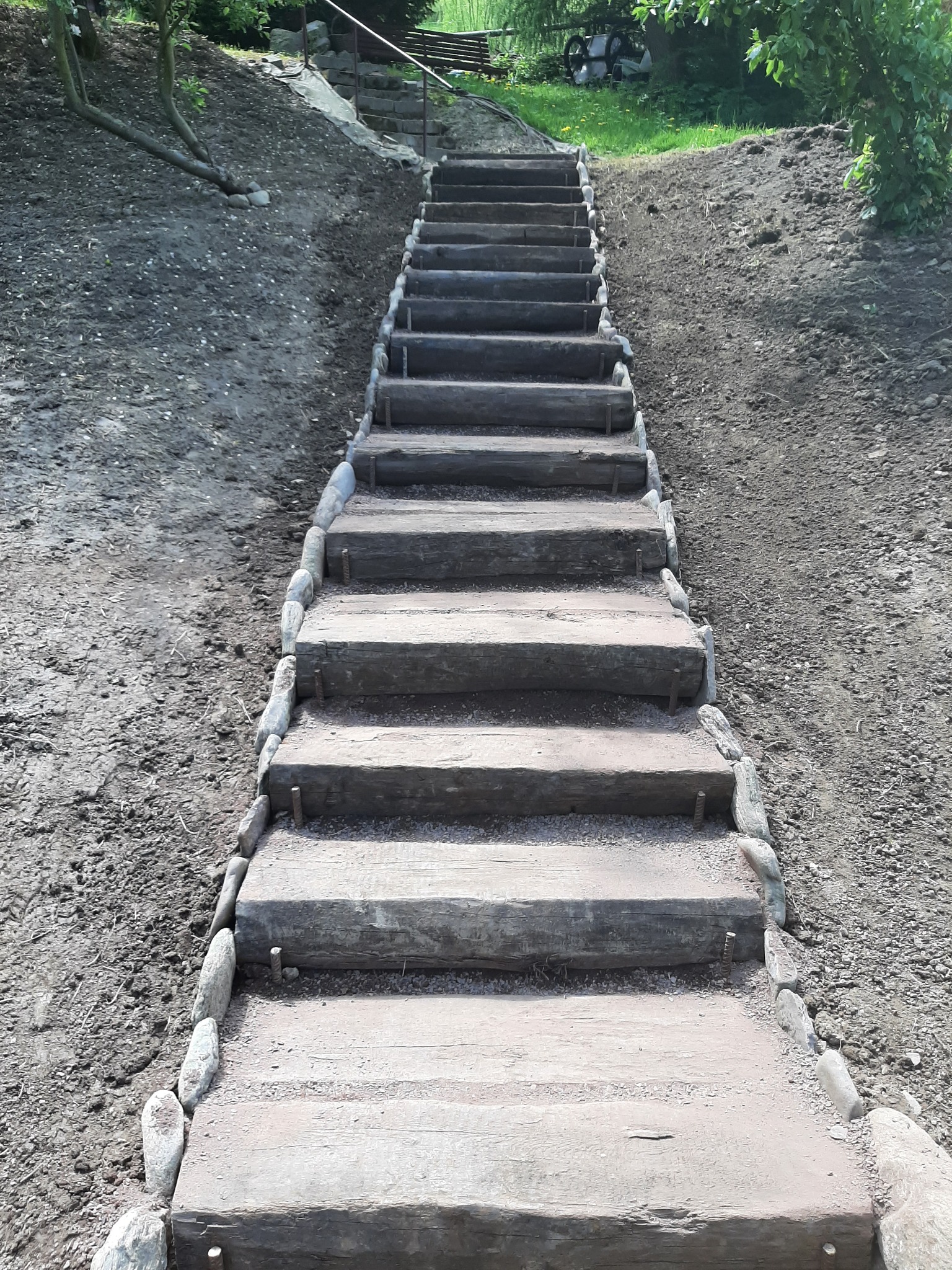 Drewniane schody z kamiennymi obrzeżami prowadzące w górę zbocza, świeżo obsypane ziemią.