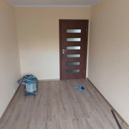 Usługi Wykończeniowo-Budowlane Neo - Pokój po remoncie z nową podłogą z paneli, widoczny odkurzacz przemysłowy, listwa przypodłogowa i niebieska zmiotka, ściany pomalowane na jasny beż.