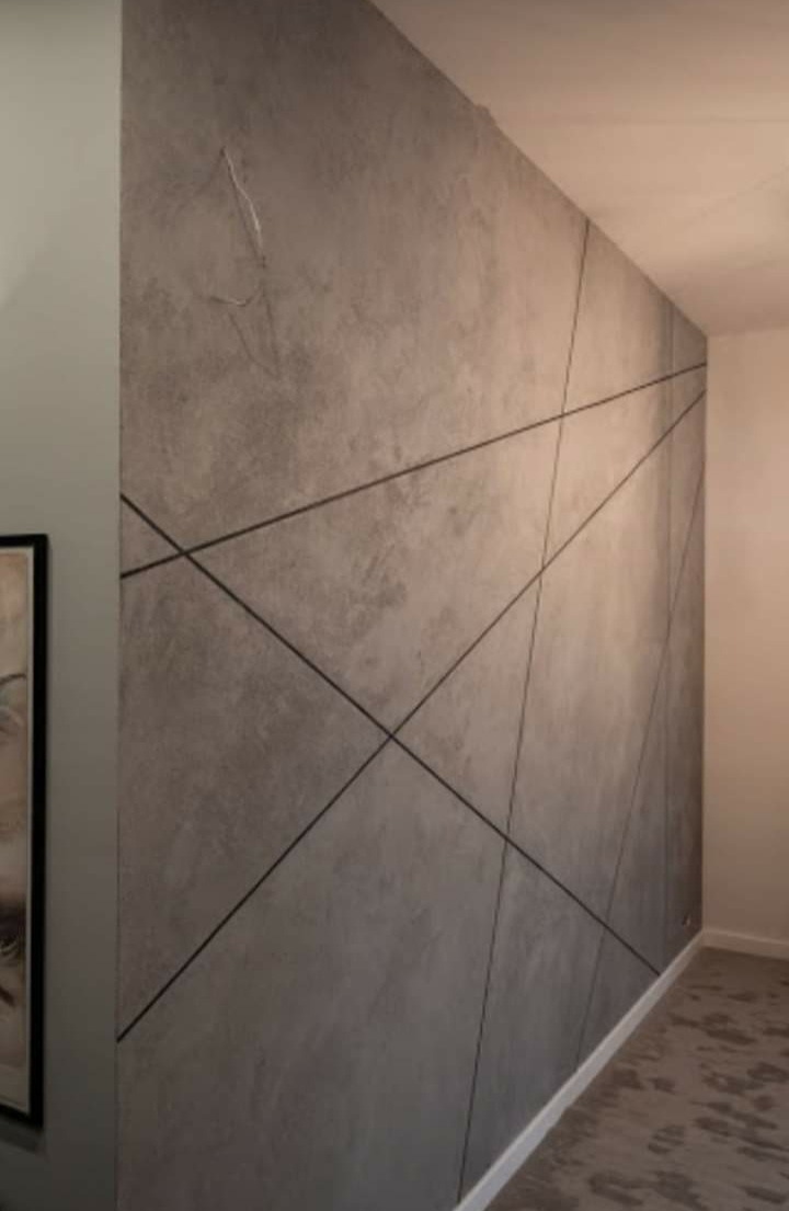 Ściana pokryta szarą, dekoracyjną fakturą z geometrycznymi, czarnymi liniami, widoczny fragment obrazu w ramie i dywan z wzorem.