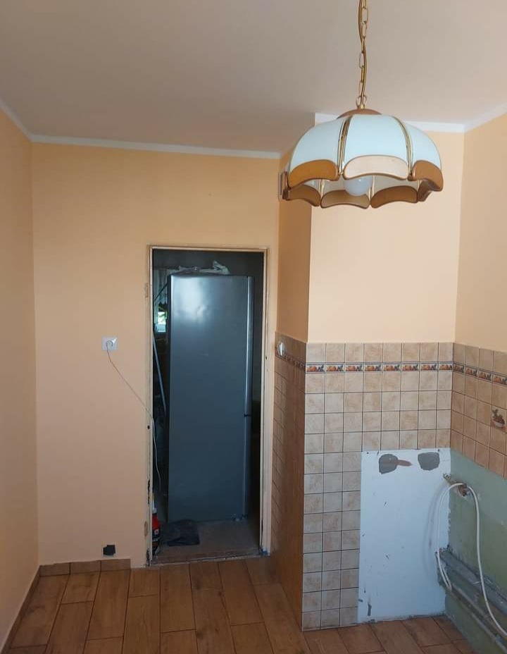 Kuchnia w trakcie remontu: położona podłoga z paneli imitujących drewno, pomalowane na brzoskwiniowo ściany, częściowo zdjęte płytki ścienne, widoczny stary żyrandol.