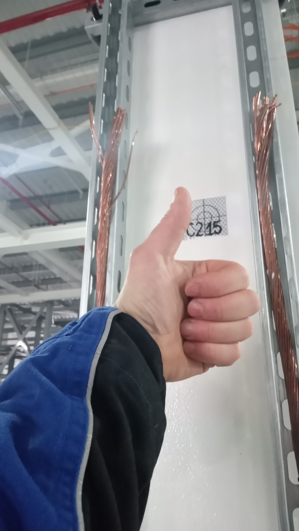 Kciuk w górę na tle metalowej konstrukcji z kablami miedzianymi i naklejką C215, sugerujący akceptację wykonanej pracy.
