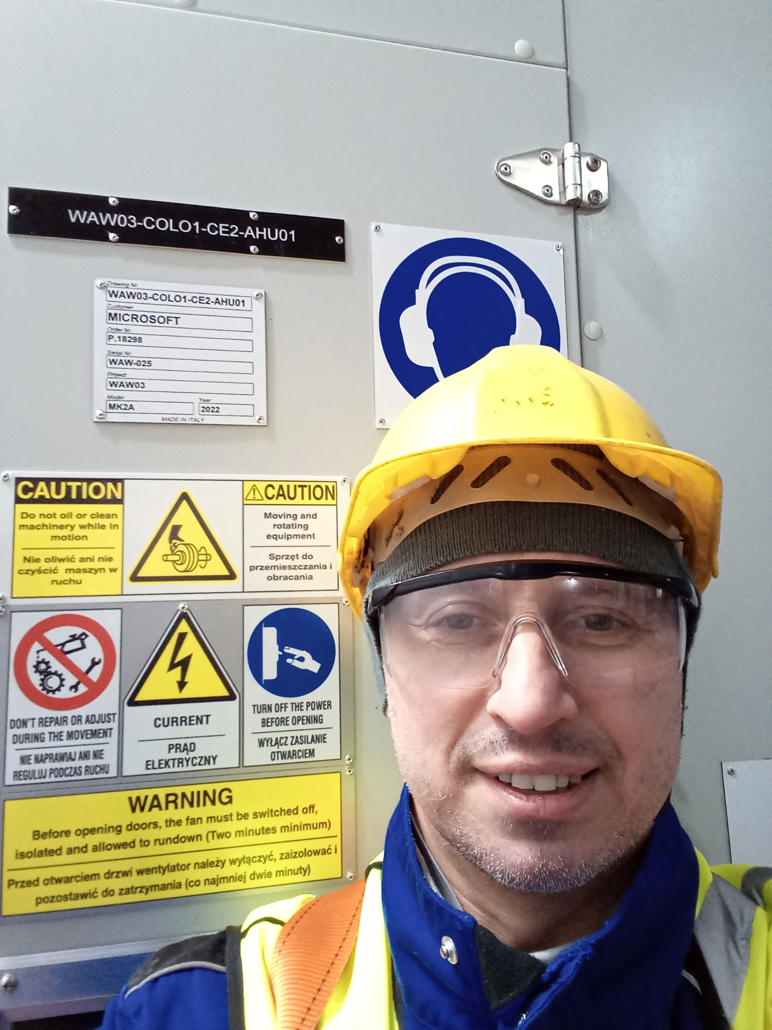 Pracownik w kasku i okularach ochronnych stoi przed szafą sterowniczą z oznaczeniami ostrzegawczymi, w tle widoczna tabliczka z napisem WAW03-COLO1-CE2-AHU01 oraz etykieta MICROSOFT.