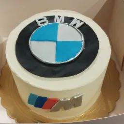 Tort urodzinowy z logo BMW i literą M w kolorach niebieskim, granatowym i czerwonym, umieszczony w kartonowym pudełku.