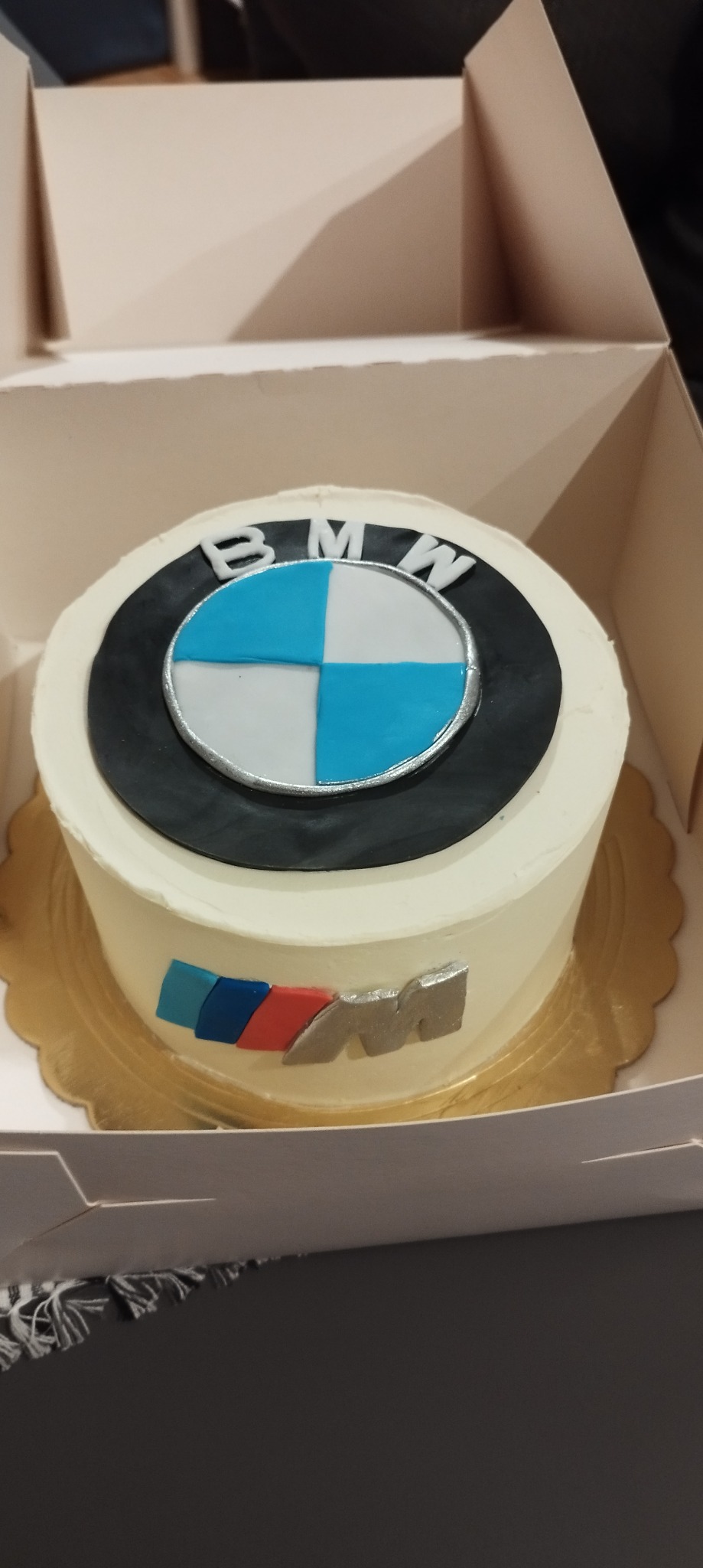 Tort urodzinowy z logo BMW i literą M w kolorach niebieskim, granatowym i czerwonym, umieszczony w kartonowym pudełku.