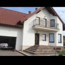 Biały dom jednorodzinny z czerwonym dachem i balkonem z czarną balustradą, widok z zewnątrz, schody wejściowe wyłożone kostką.