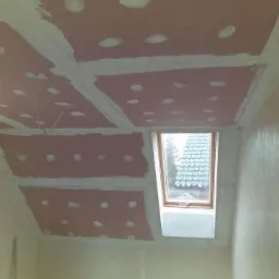 Montaż płyt gipsowo-kartonowych na poddaszu ze skosami, widoczne łączenia szpachlowane na biało, okno dachowe w tle.