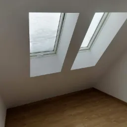 Poddasze z dwoma oknami dachowymi, przez które widać padający śnieg, świeżo pomalowane ściany na biało i drewniana podłoga.