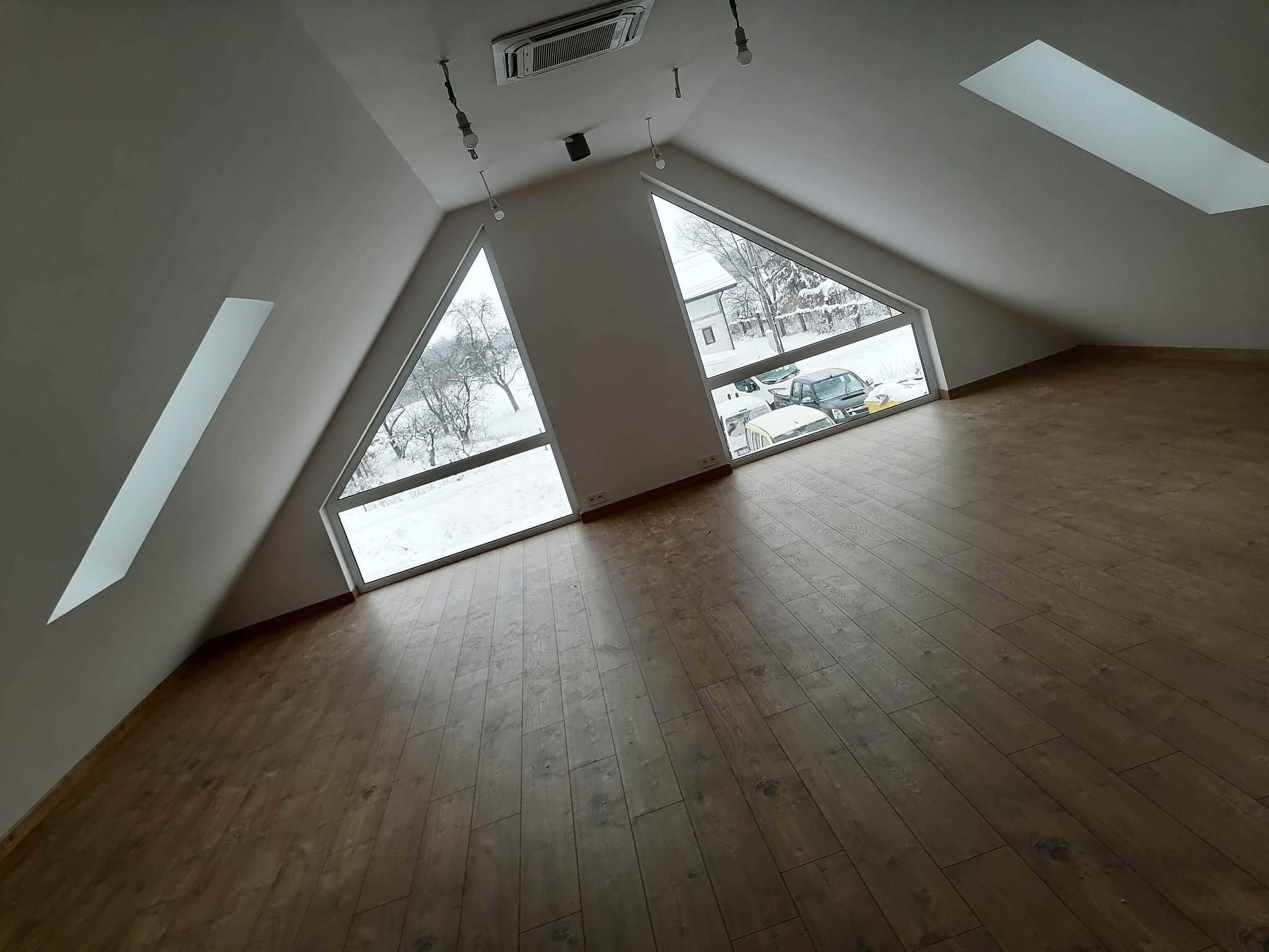Puste poddasze z drewnianą podłogą, skośnymi ścianami, oknami trójkątnymi i lukarnami, widok na zaśnieżone drzewa i samochody na zewnątrz.