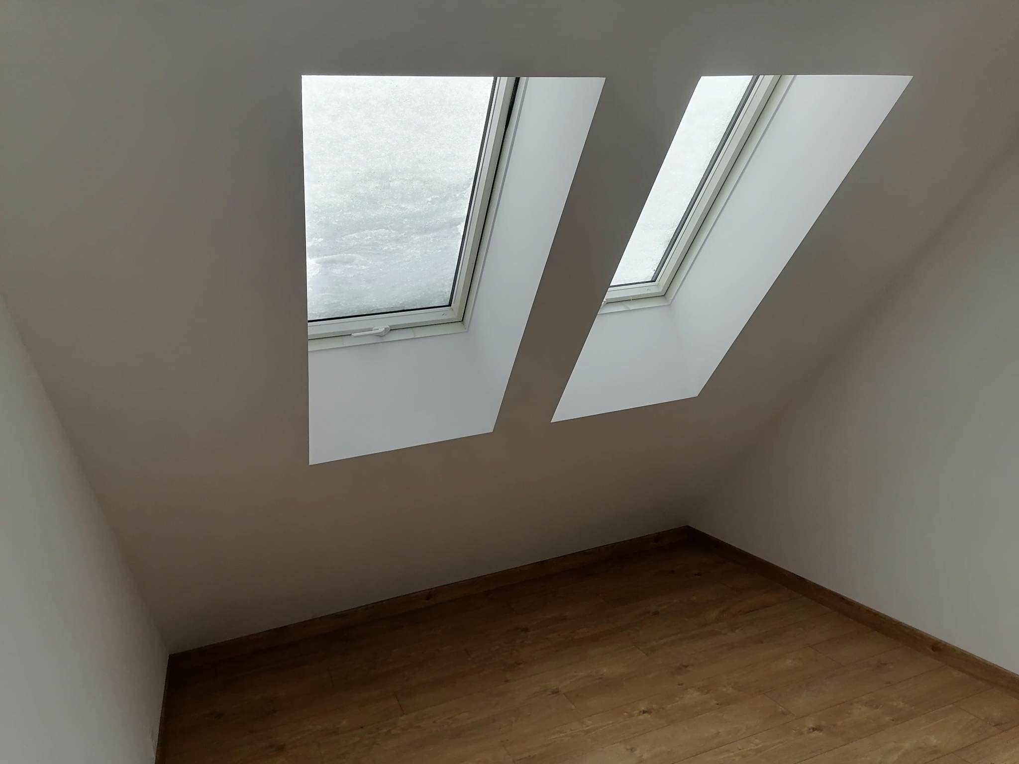 Poddasze z dwoma oknami dachowymi, przez które widać padający śnieg, świeżo pomalowane ściany na biało i drewniana podłoga.