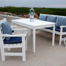 Biały zestaw mebli ogrodowych: stół, ławka z poduszkami w paski i krzesło z granatową poduszką, uzupełnione dekoracyjną latarenką z numerem 1890, na tle kamiennego brzegu i zieleni.