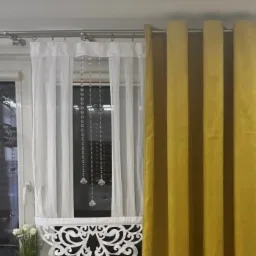 Okno z białą, prześwitującą firanką z dekoracyjnym, ażurowym wykończeniem i kryształkami, obok zasłona w kolorze musztardowym, na parapecie wazony z kwiatami.