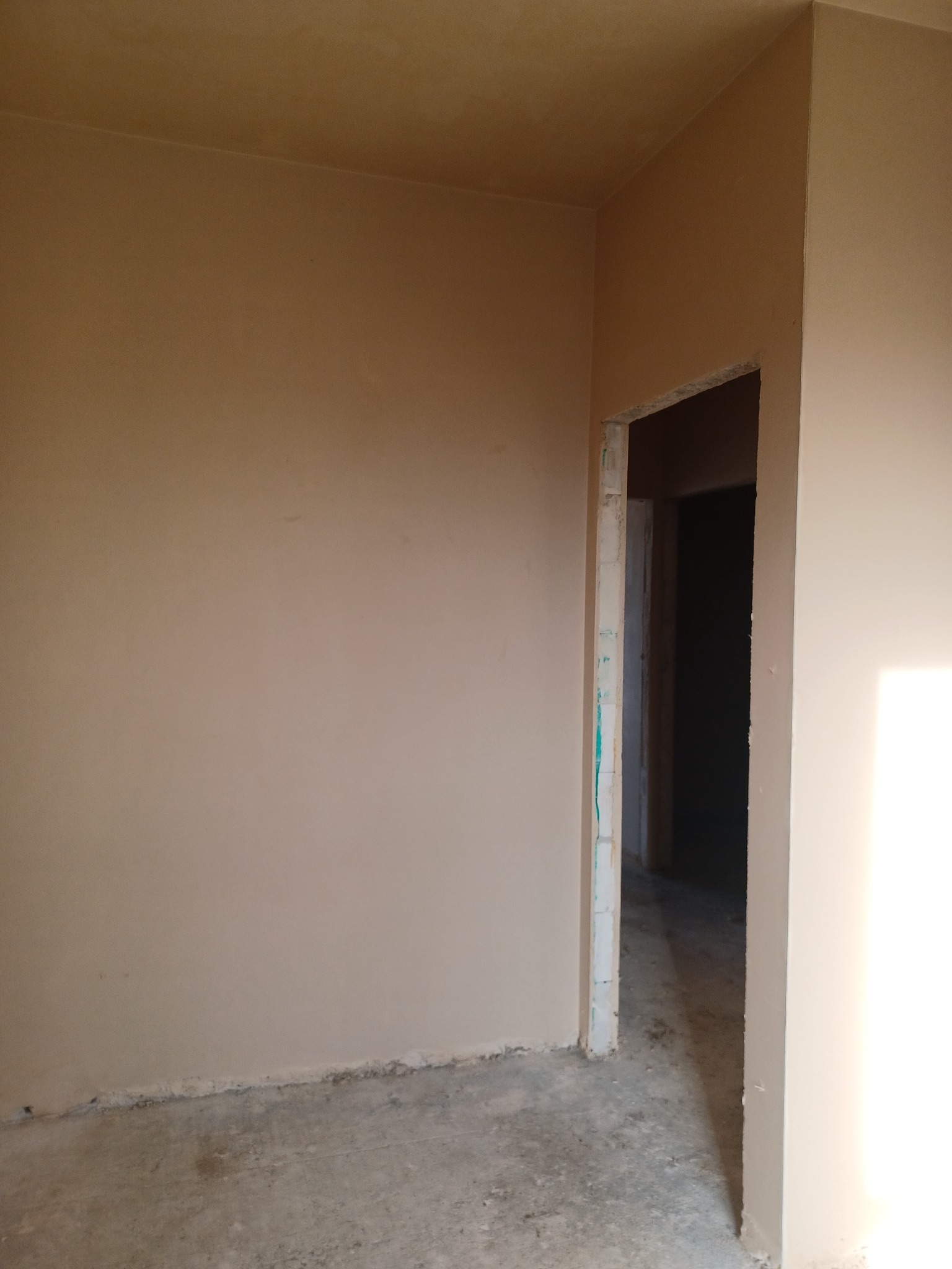 Surowe wnętrze w trakcie prac wykończeniowych: świeżo otynkowane ściany w beżowym odcieniu, widoczny surowy beton na podłodze i ościeżnica drzwiowa z resztkami zaprawy, czekające na dalsze prace.