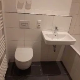 Nowoczesna łazienka po remoncie: biała umywalka, podwieszana toaleta, ciemnoszara podłoga z płytek, grzejnik drabinkowy i białe płytki ścienne z chromowanymi listwami.